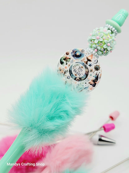 Decorative broom with teal handle and pink feathers on a white background, featuring 'Mandy's Crafting Shop' branding.